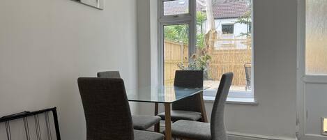 A dining area with a glass table and four grey upholstered chairs. A large abstract painting is on the wall, and a window provides a view of a backyard with a fence and greenery.