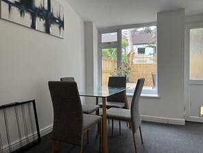 A dining area with a glass table and four grey upholstered chairs. A large abstract painting is on the wall, and a window provides a view of a backyard with a fence and greenery.