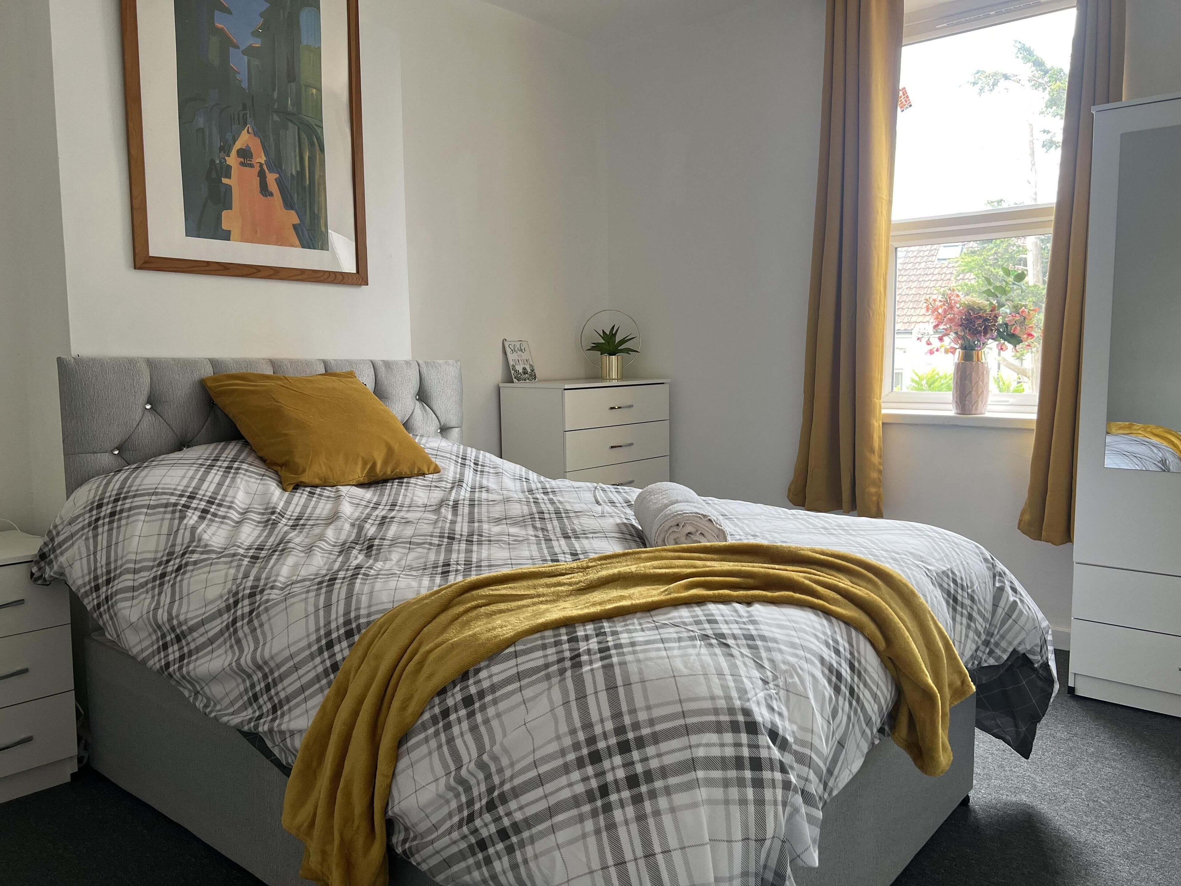 A bedroom with a bed, a bedside table, a nightstand, a window with curtains, and a painting on the wall.