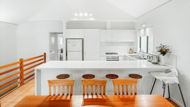 A modern kitchen with white cabinets, a central island with bar stools, and a wooden dining table.
