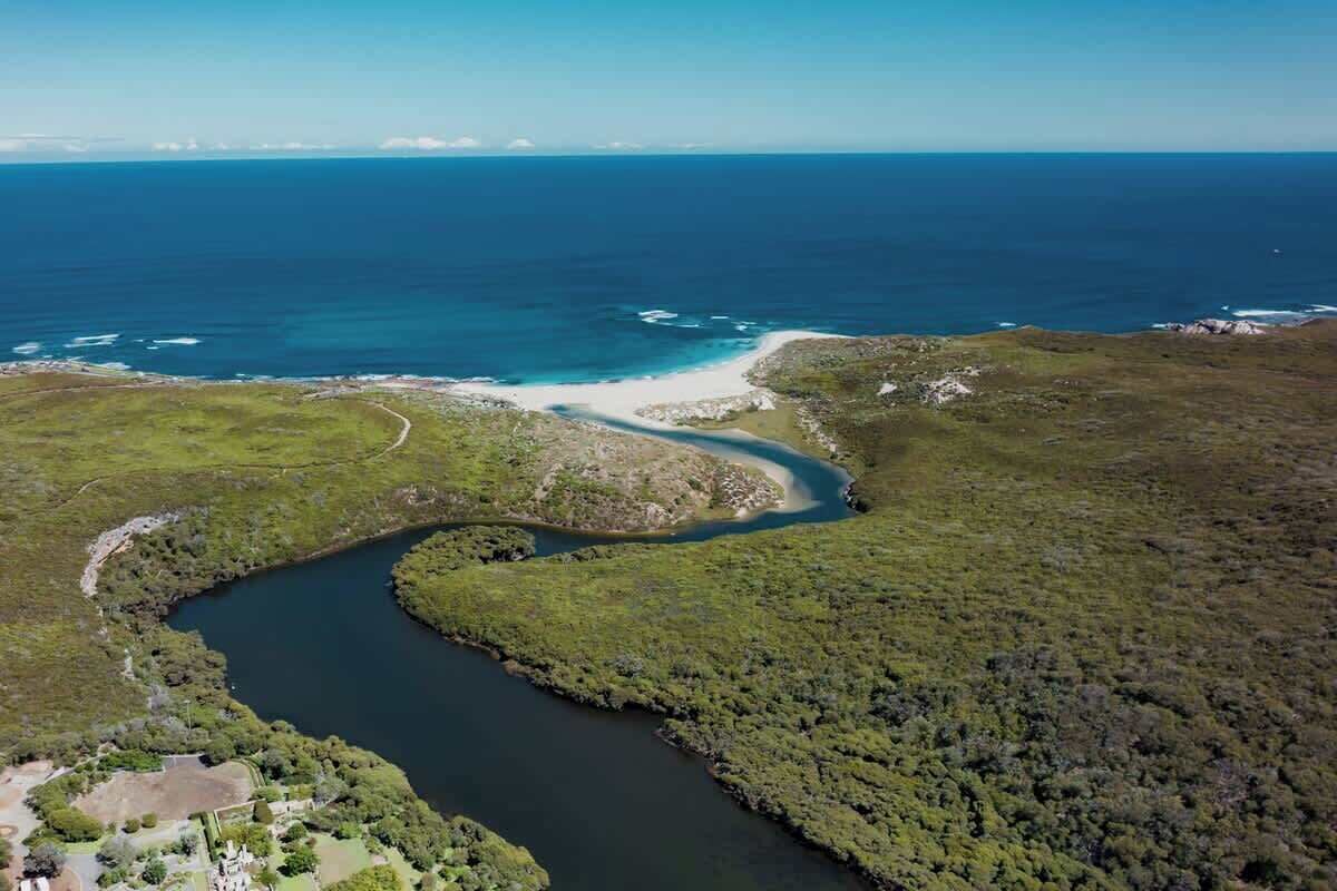 An aerial view of a coastal area with a winding river, a beach, and a small settlement with buildings and greenery.
