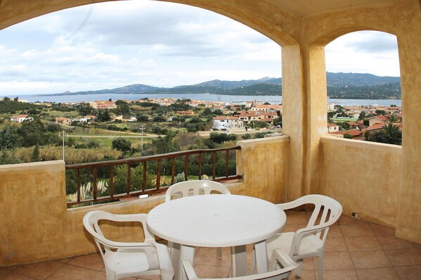 Un balcón con una mesa y sillas, con vista a un pueblo costero con edificios y montañas a lo lejos.