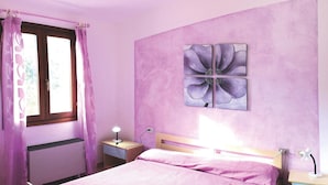 Ein Schlafzimmer mit Bett, Nachttisch, Schreibtisch, einem Fenster mit Vorhängen und einem Blumenbild an der Wand.