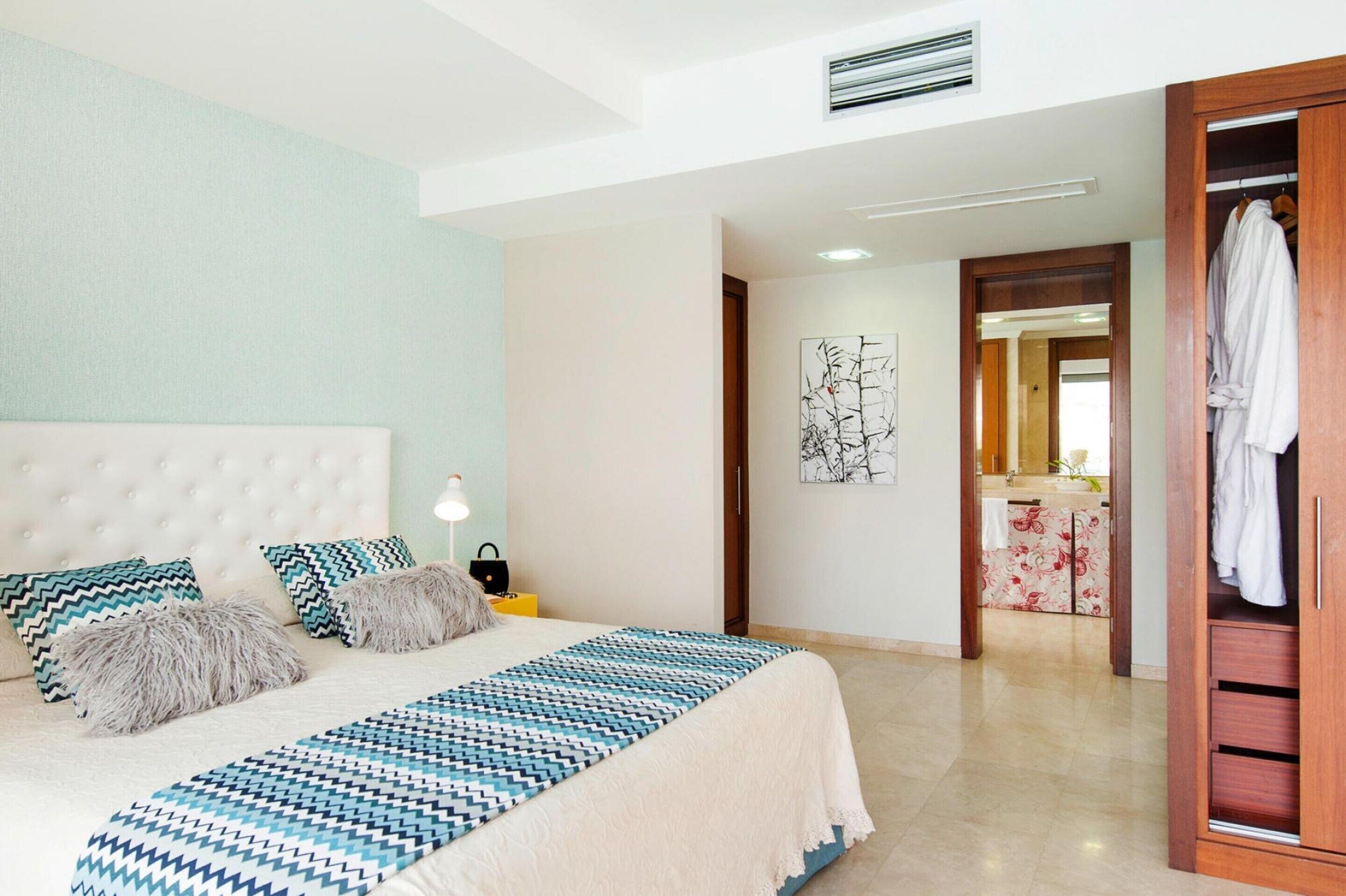Un dormitorio con una cama grande, una mesita de noche con lámpara, un armario y un baño visible a través de una puerta abierta.
