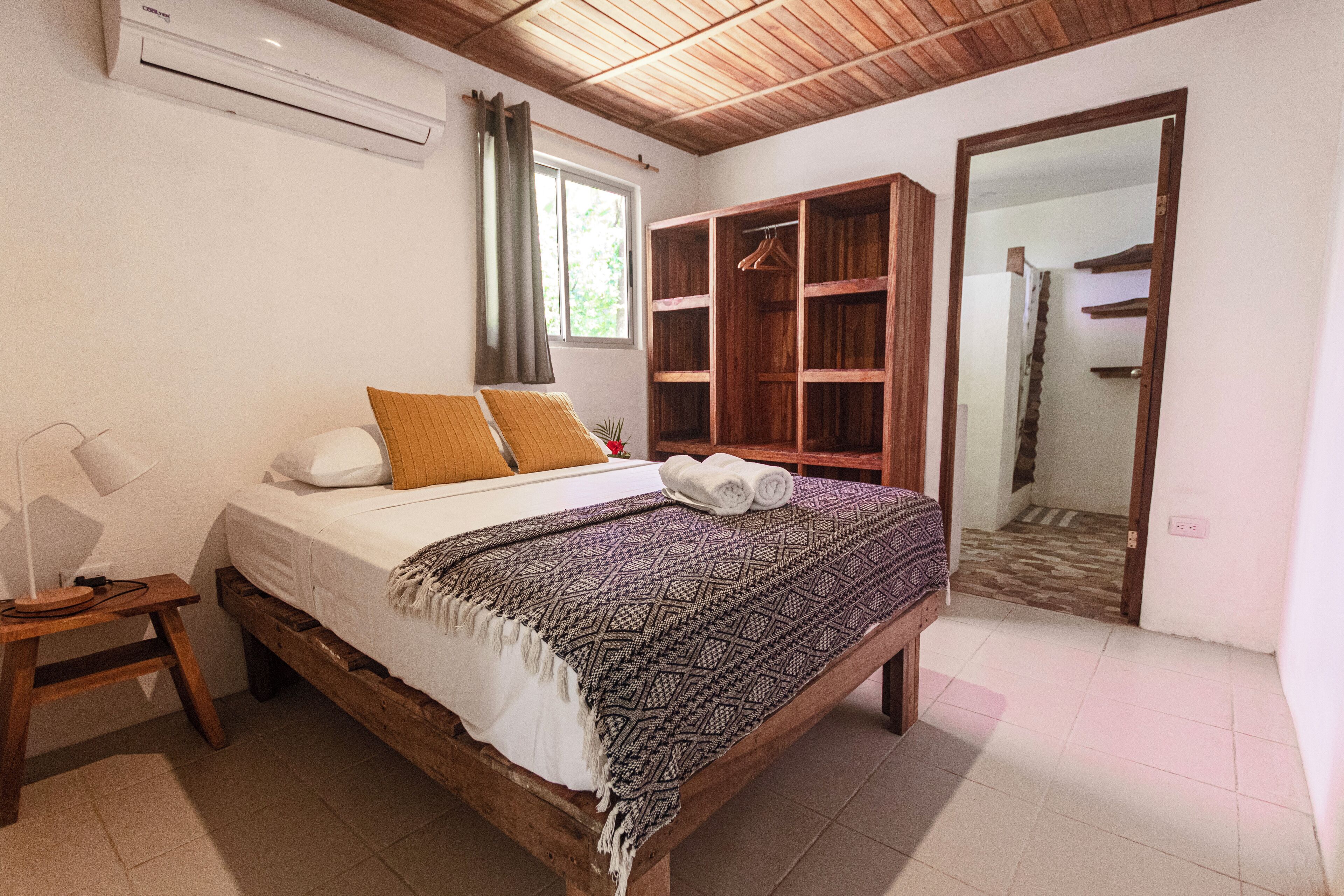Un dormitorio con cama de madera, una mesita de noche con lámpara, un armario y un baño visible a través de una puerta abierta.