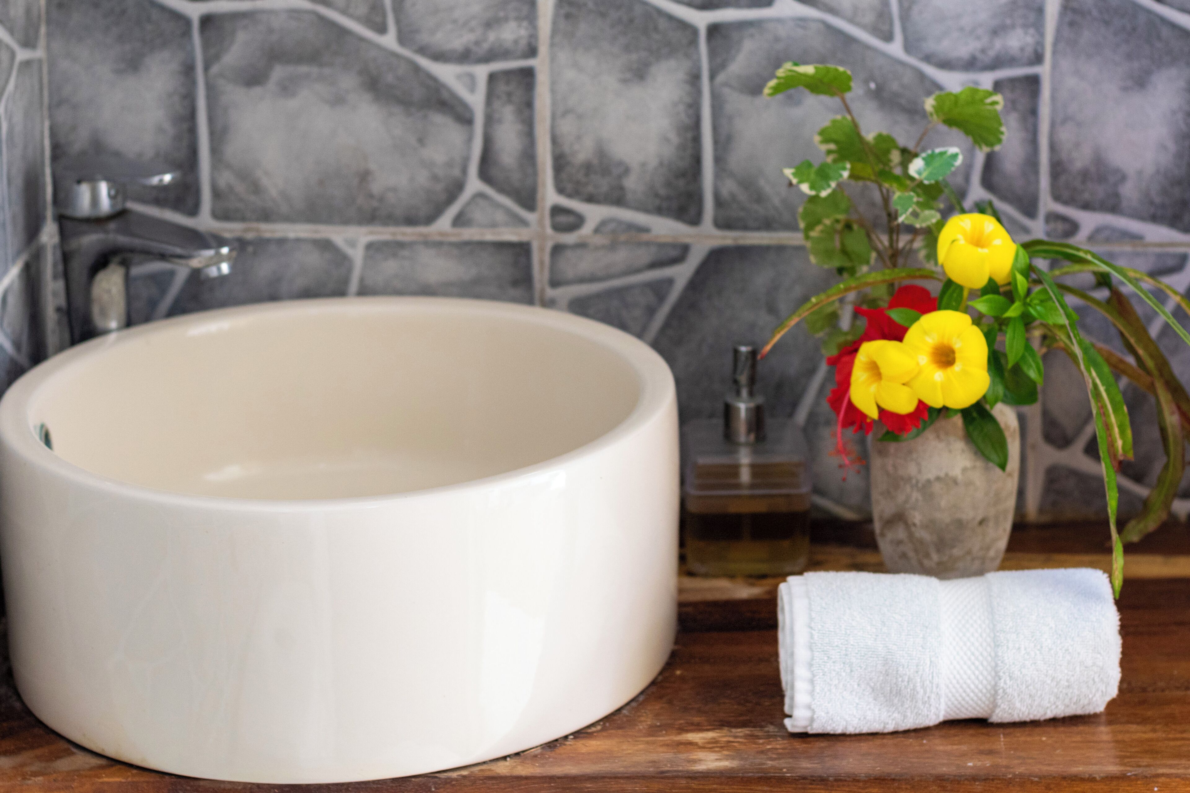 A bathroom with a round white sink, a stone wall, and a vase with yellow flowers.