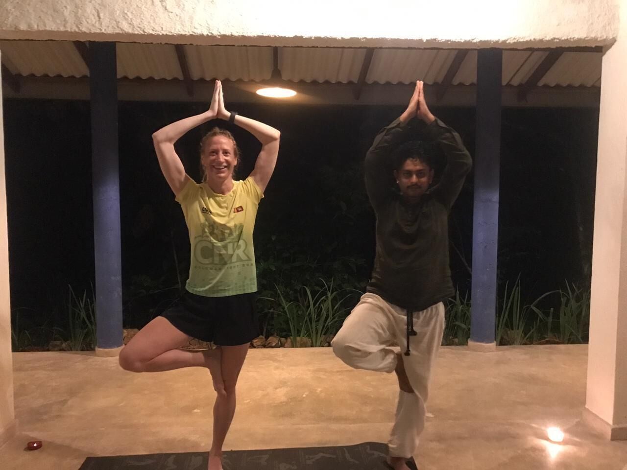 Deux personnes pratiquent des postures de yoga à l’extérieur, dans un espace abrité doté d’un sol en béton et agrémenté de plantes en arrière-plan.