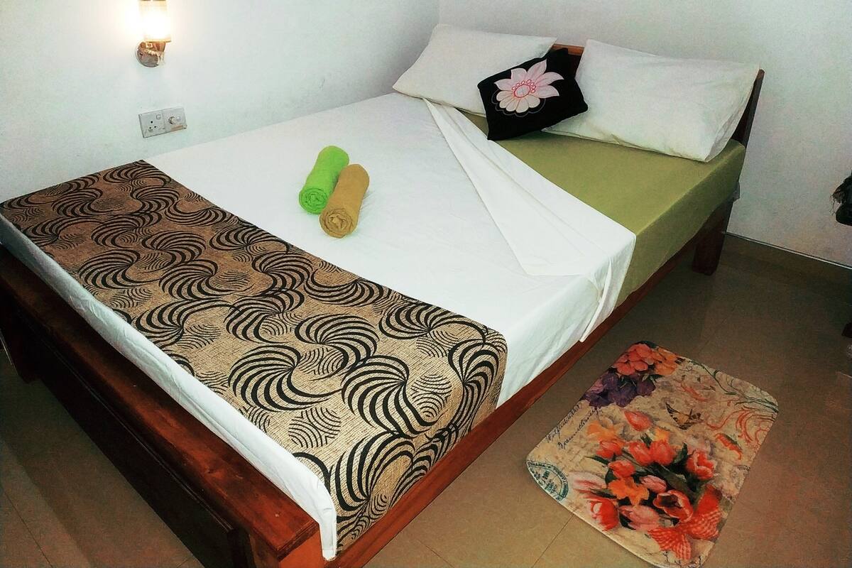 Ein Einzelbett mit gemustertem Bettbezug, zwei zusammengerollte Handtücher, ein Kissen mit Blumenmuster und ein floraler Teppich auf dem Boden.