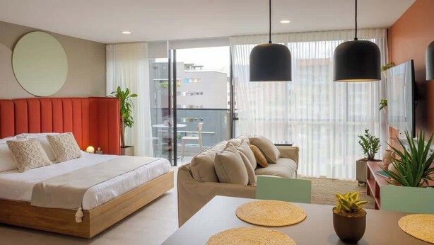 Moderni hotellihuone, jossa on sänky, sohva, ruokapöytä ja parveke, jolta on näkymä.