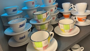 Diverse witte keramische kopjes en schoteltjes met verschillende designs en logo's, uitgestald op een plank.