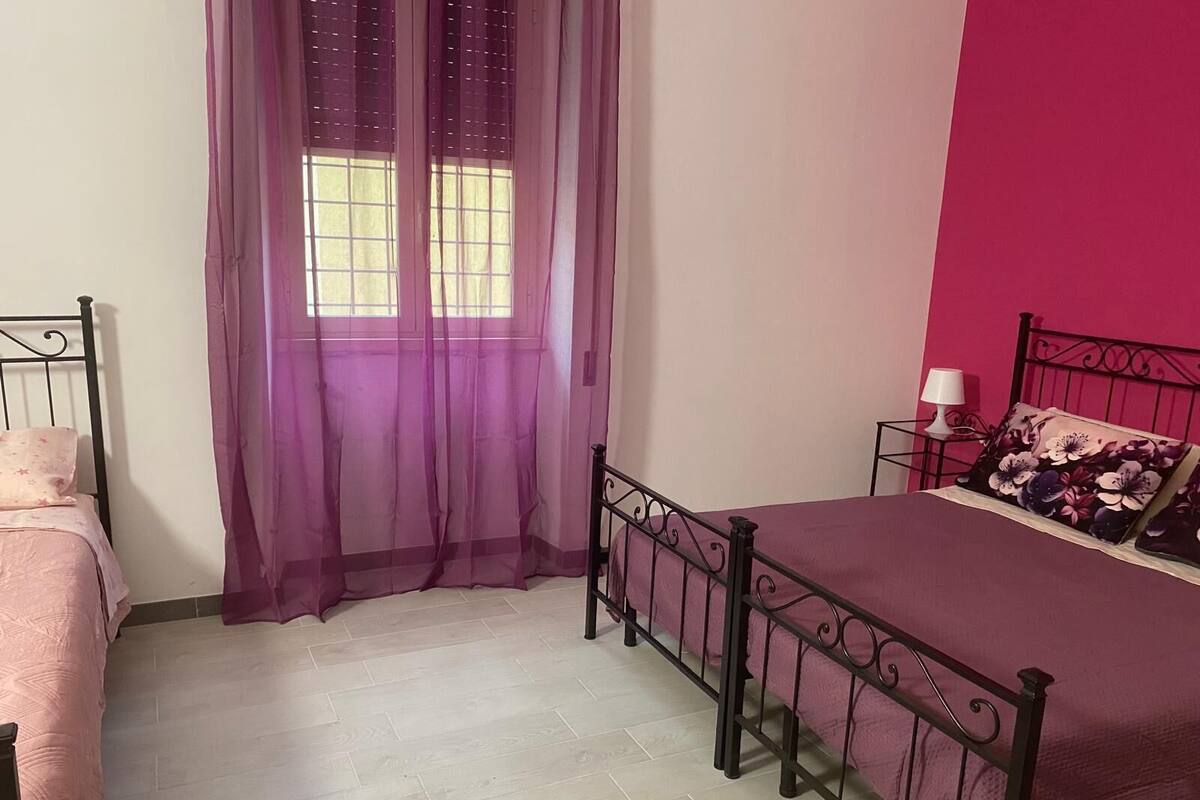 A bedroom with a bed, a window with purple curtains, a nightstand with a lamp, and a pink wall.