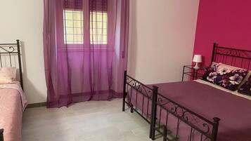 A bedroom with a bed, a window with purple curtains, a nightstand with a lamp, and a pink wall.