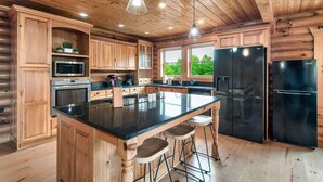 A spacious kitchen with wooden cabinets, a large island with stools, stainless steel appliances, and a view of greenery outside.