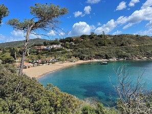 Ett kustområde med en sandstrand, klart blått vatten och en sluttning med byggnader.
