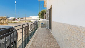 Un balcón con piso de baldosas, baranda de metal y vista a una calle con edificios y cielo despejado.
