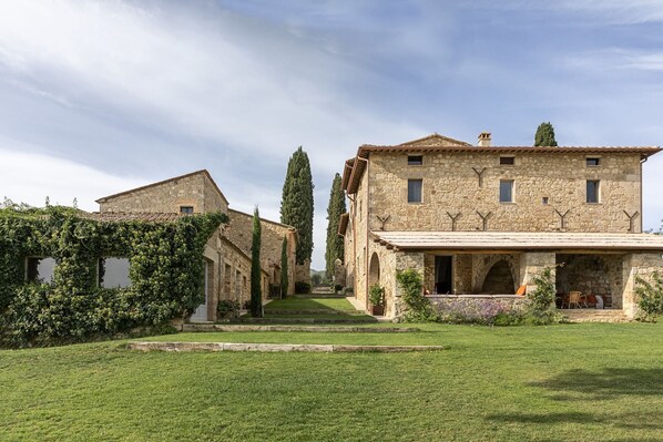 Traditional stone building with a central entrance and covered porch, surrounded by lush greenery and tall cypress trees.