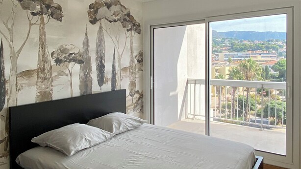 A bedroom with a large bed, a view of a balcony, and a cityscape through the window.