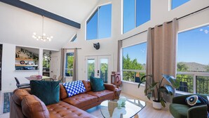 A spacious living room with a brown leather sectional sofa, a glass coffee table, and a green armchair. There is a balcony with a view of greenery and a dining area with a table and chairs. - Stylish Lake Travis Getaway! Beach Access, Poker & VR (Lago Vista)