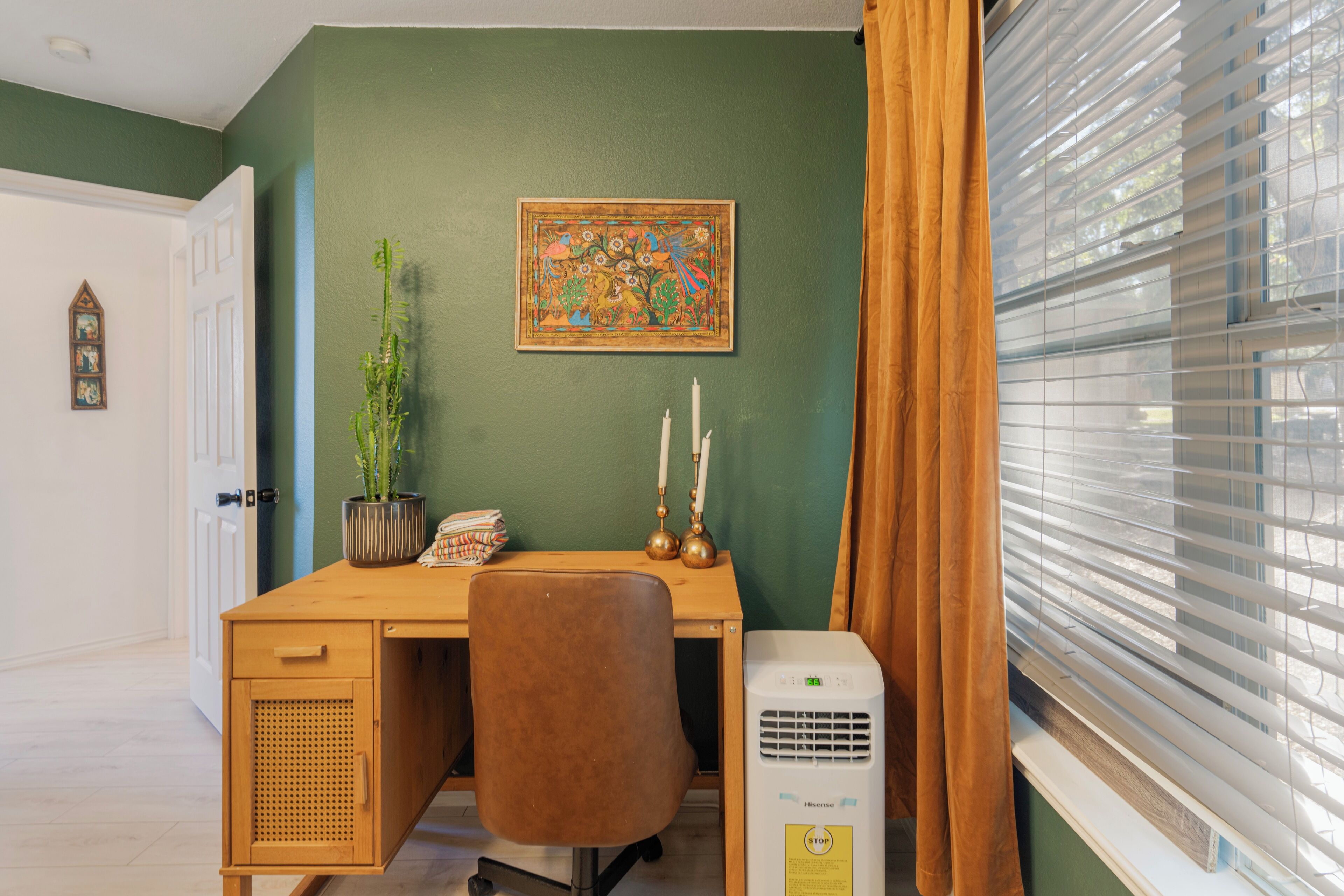 A room with a desk, chair, potted plant, wall art, candles, and a window with blinds.