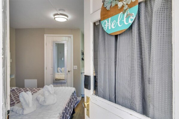 Une chambre d’hôtel avec un lit, une porte sur laquelle est affiché le mot « Bonjour », et une fenêtre avec des rideaux à motifs.