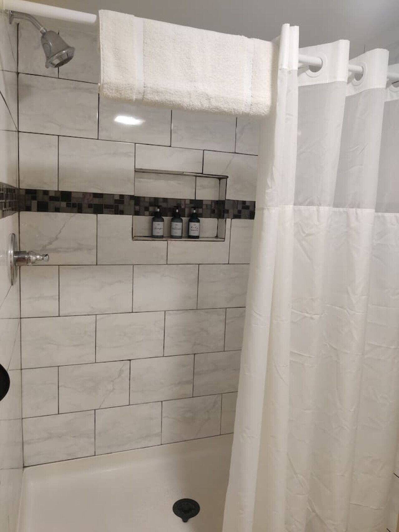 A shower area with a tiled wall, a shelf with bottles, and a white shower curtain.