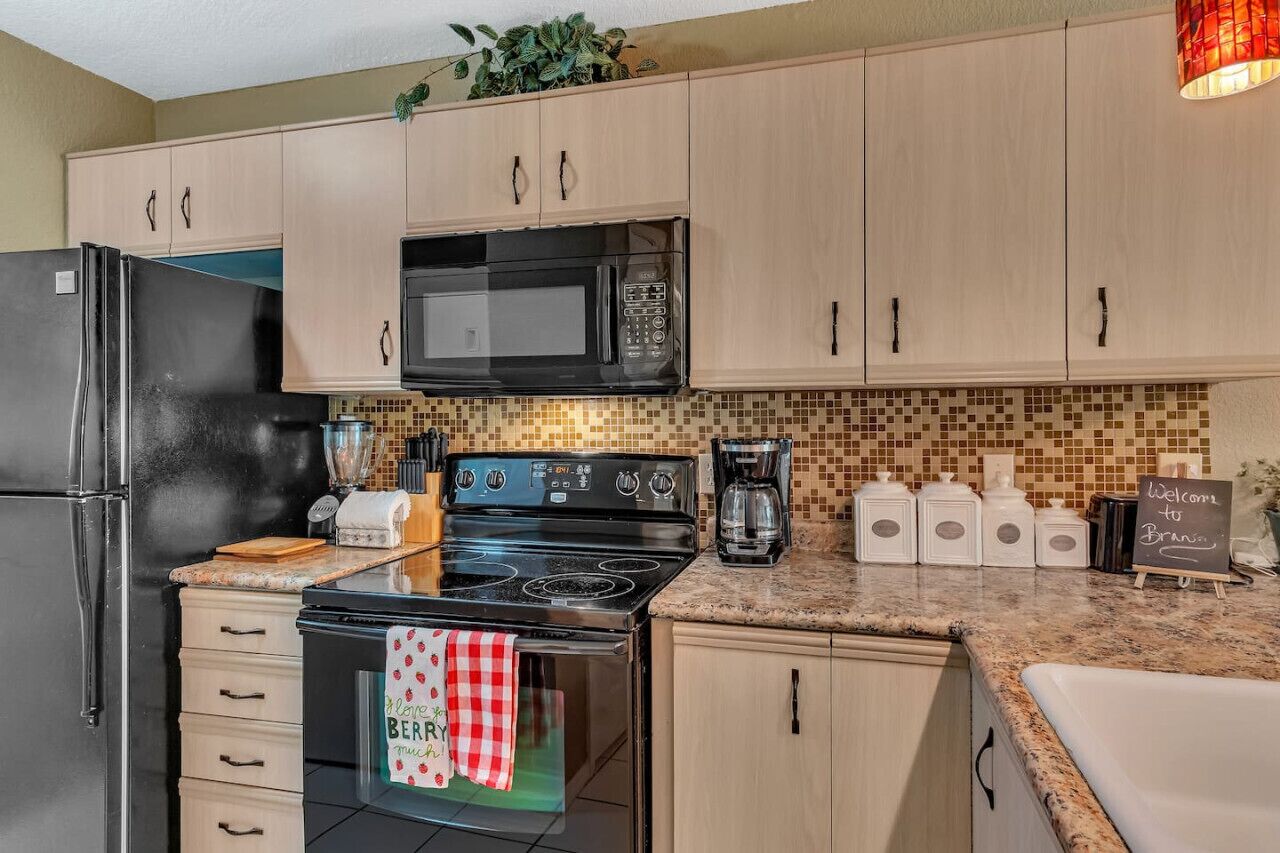 A kitchen with light wooden cabinets, a black refrigerator, a microwave, a stove, and a coffee maker. There is a chalkboard sign that says "Welcome to Braun."