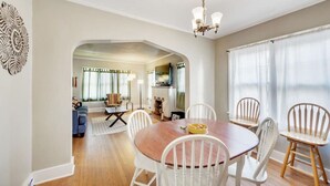 A dining area with a wooden table and white chairs, a chandelier, and a view into the living room. - Golden Luxe Bungalow | Walk to Downtown & Parks (Golden)