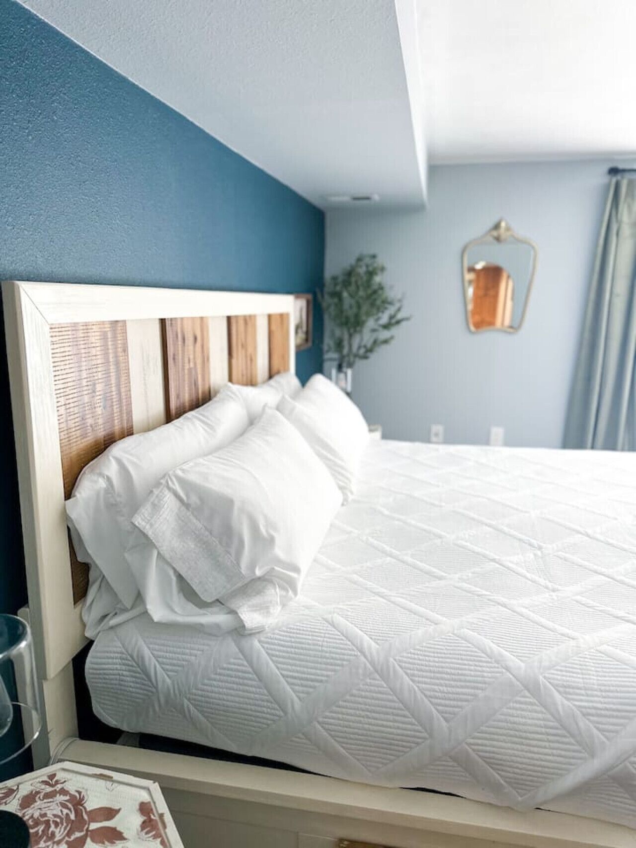 Una cama bien tendida con ropa blanca y cabecero de madera en una habitación con paredes azules y una planta pequeña al lado.