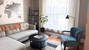 Una sala de estar moderna con un sofá gris, una mesa de centro redonda negra y un sillón azul. Hay un mapa del mundo en la pared, una estantería y una planta en maceta.
