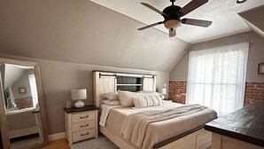 Un dormitorio con cama, mesitas de noche, cómoda, ventilador de techo y ventana con cortinas.