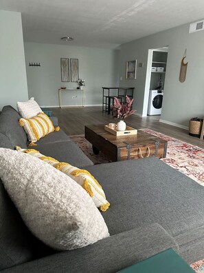 Een moderne woonkamer met een grijze bank, een houten salontafel en een vloerkleed met een patroon.