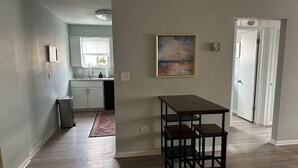 A spacious room with a wooden floor, a dining table with chairs, a kitchen area with cabinets and a window, and a painting on the wall. - Lakewood Gem: Furnished Haven! Perfect for Month Long Stays! (Lakewood)