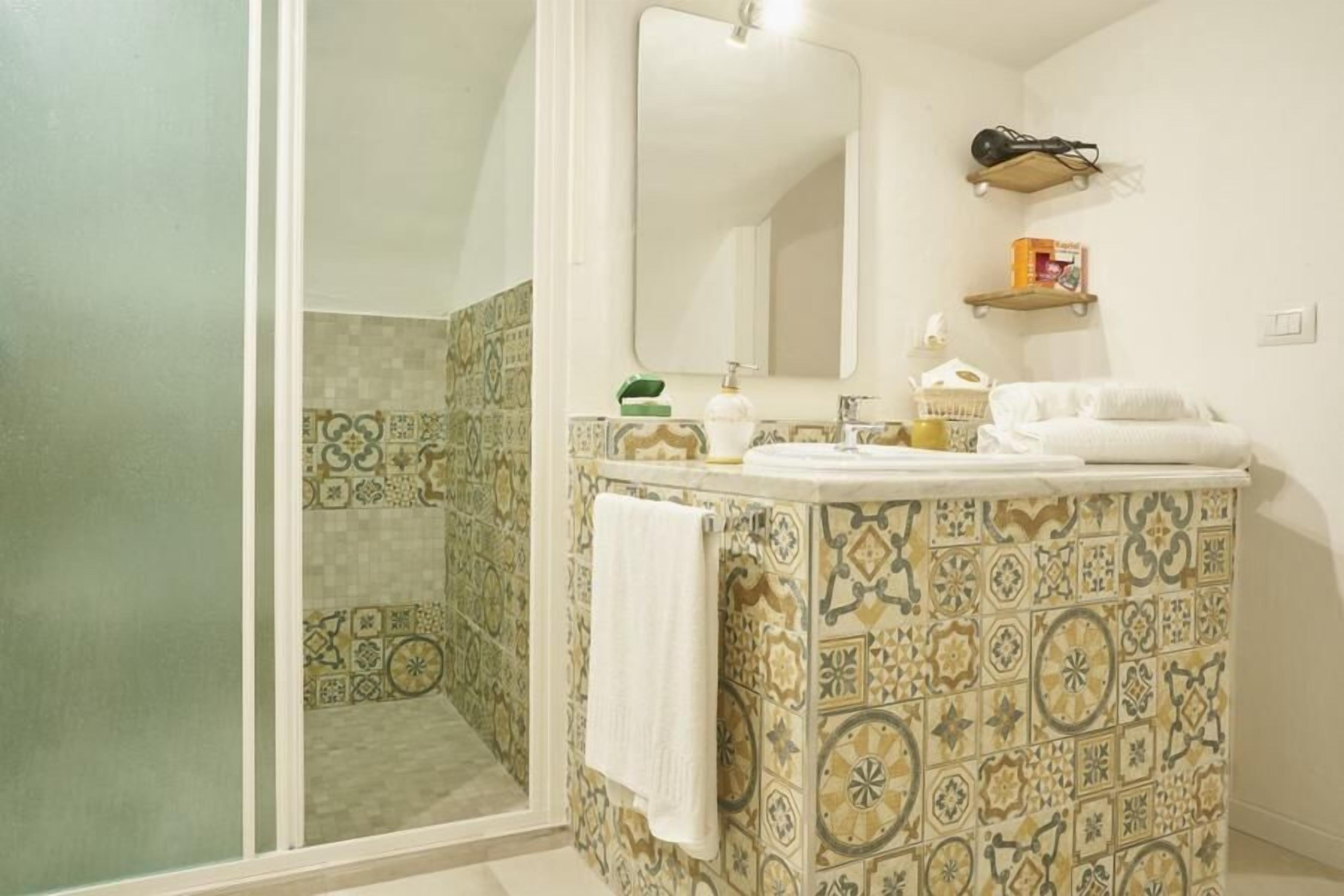 A bathroom with a tiled wall, a sink, a mirror, and a shower area.