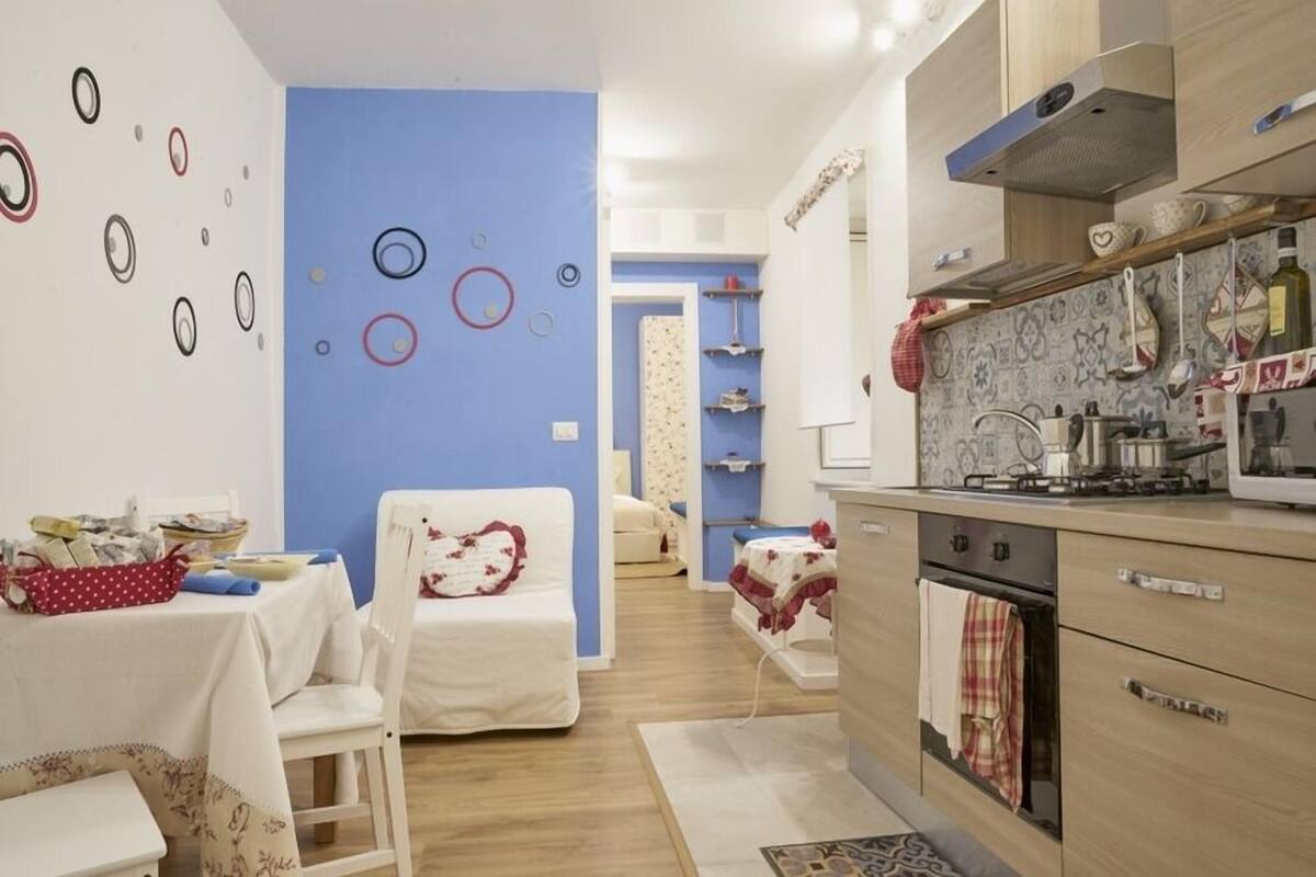 A modern kitchen with a stove, oven, and various kitchen utensils. There is a dining area with a table and chairs, and a bedroom with a bed and a chair.