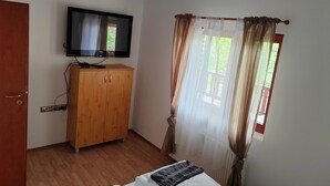 A bedroom with a wooden wardrobe, a television on the wall, a bed with a white mattress and a grey blanket, and a window with sheer curtains.