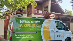 A green and white van parked in front of a building with a sign that reads "Nyiss Ajót A Kiflineik!" and "kifli.hu".