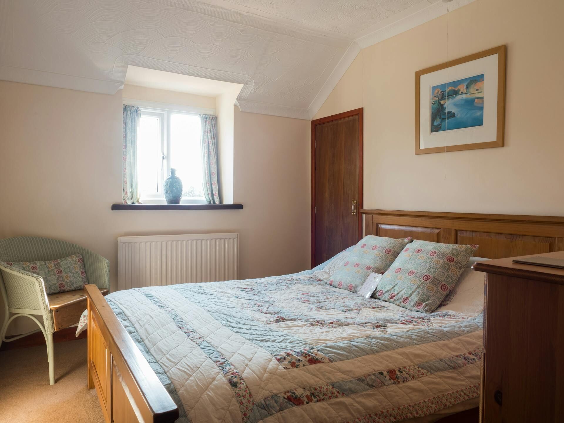 Une chambre à coucher avec un lit en bois, une fenêtre avec des rideaux, un radiateur, une chaise et un tableau accroché au mur.