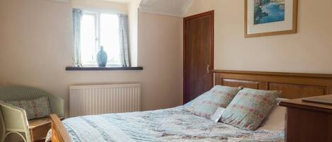 A bedroom with a wooden bed, a window with curtains, a radiator, a chair, and a painting on the wall.