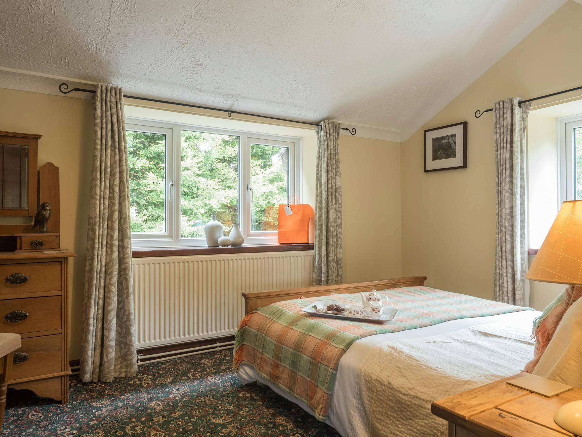 A bedroom with a bed, a dresser, a window with curtains, a radiator, and a lamp.
