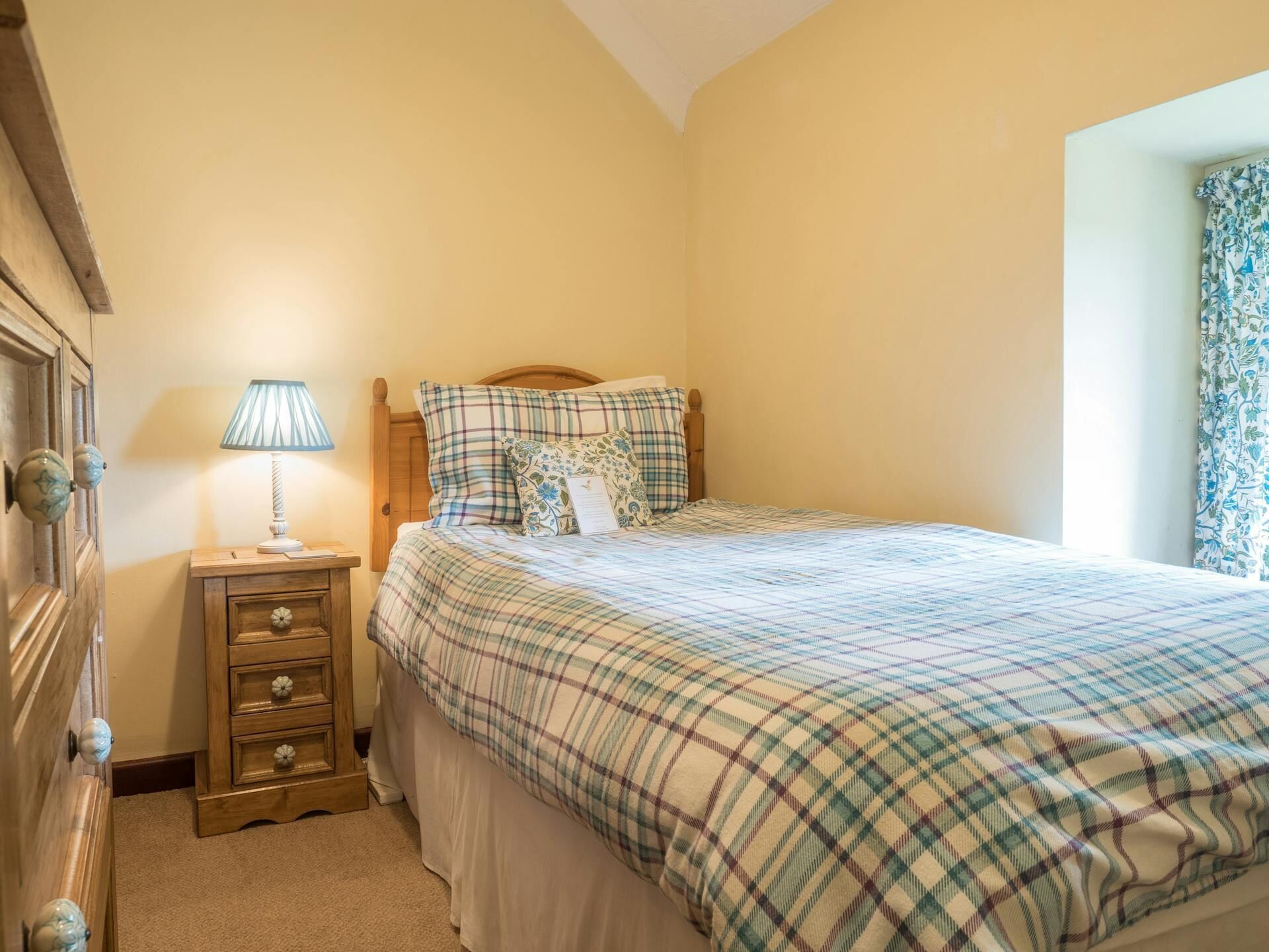 A bedroom with a bed, a nightstand with a lamp, a wooden dresser, and a window with curtains.