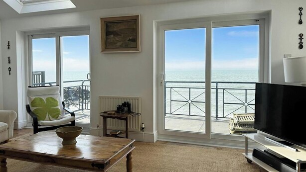 Un salon comprenant une grande télévision, une table basse, un fauteuil et un canapé. De grandes fenêtres offrent une vue sur la mer.