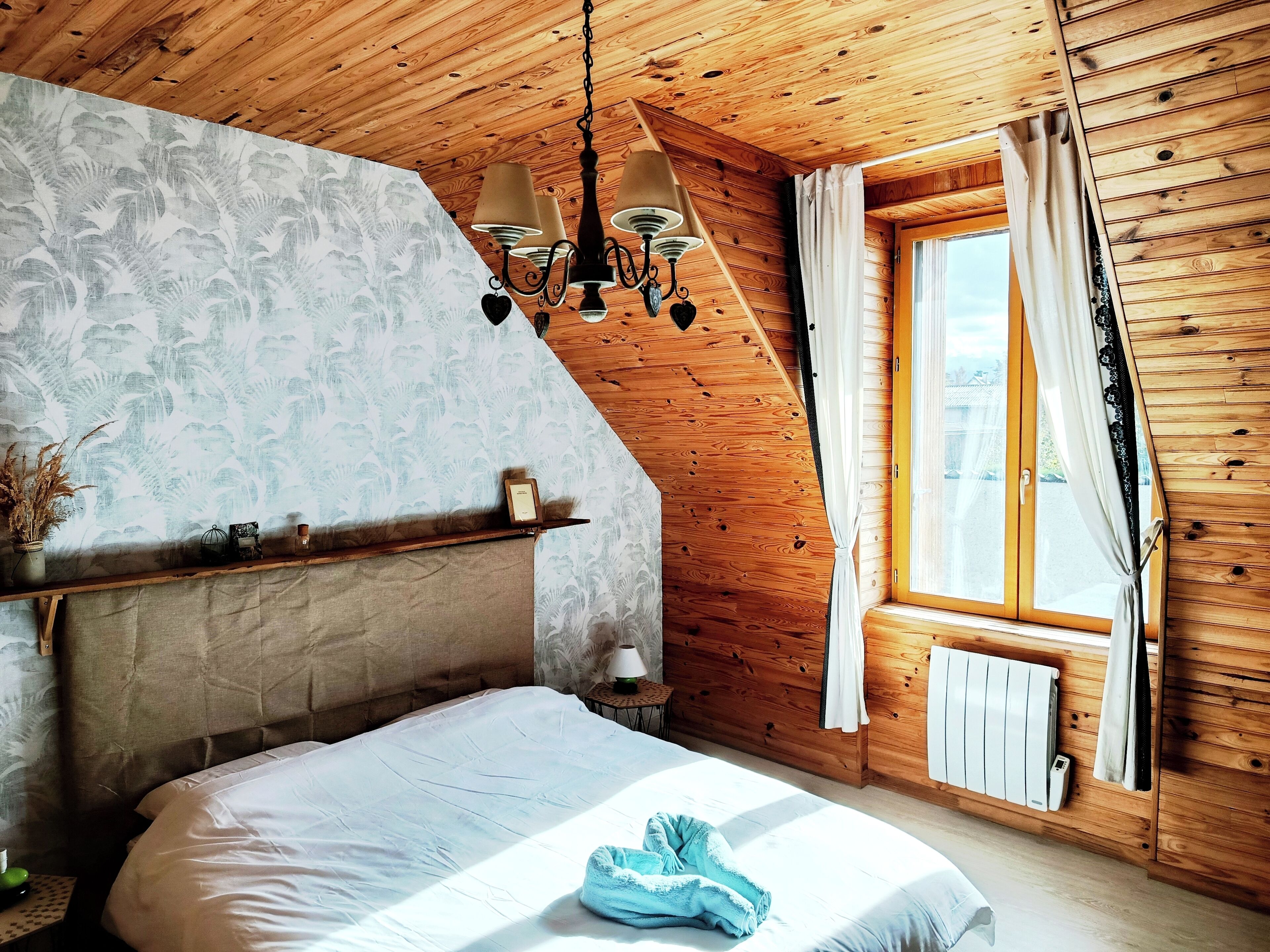 Ein Schlafzimmer mit schrägverdeckter Decke, Holzwänden, einem Bett mit weißer Bettwäsche, einem Nachttisch mit Lampe und einem Fenster mit Vorhängen.