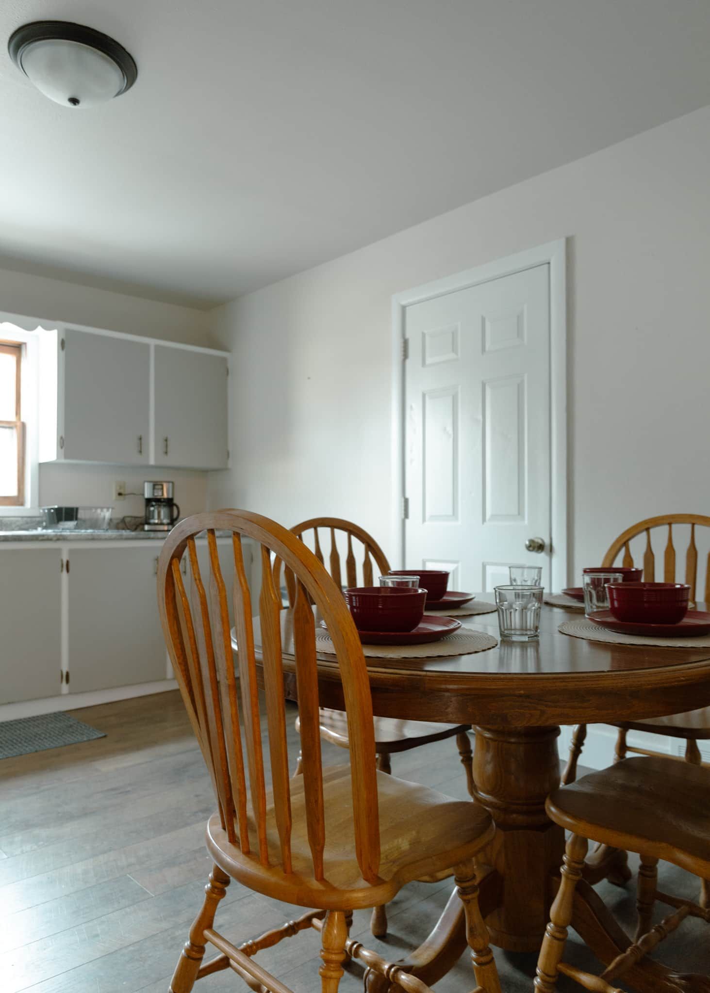 Un comedor con una mesa y sillas de madera, una puerta blanca y una cocina al fondo.