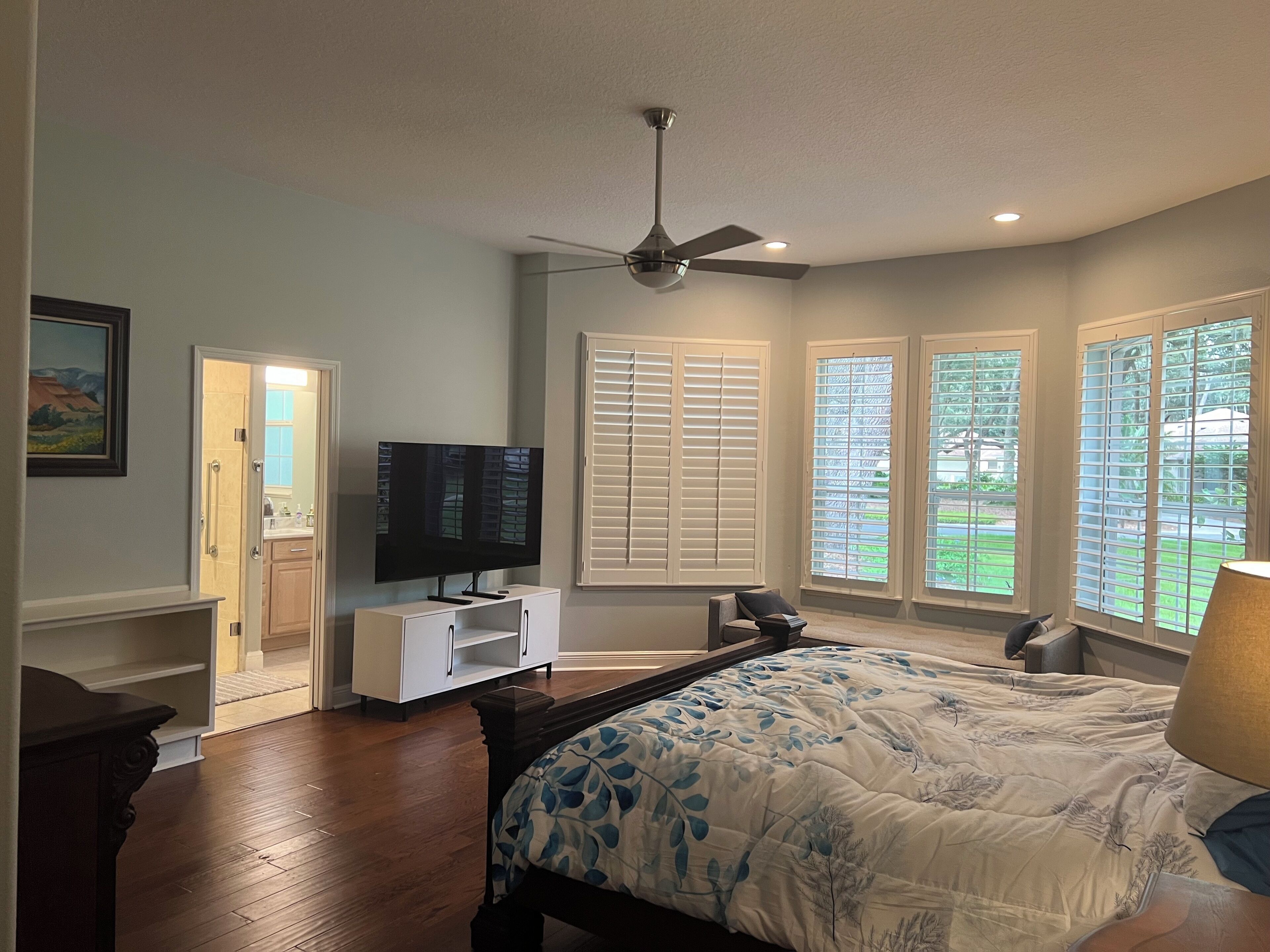 Un dormitorio con cama, televisor, ventilador de techo y ventanas amplias con persianas.