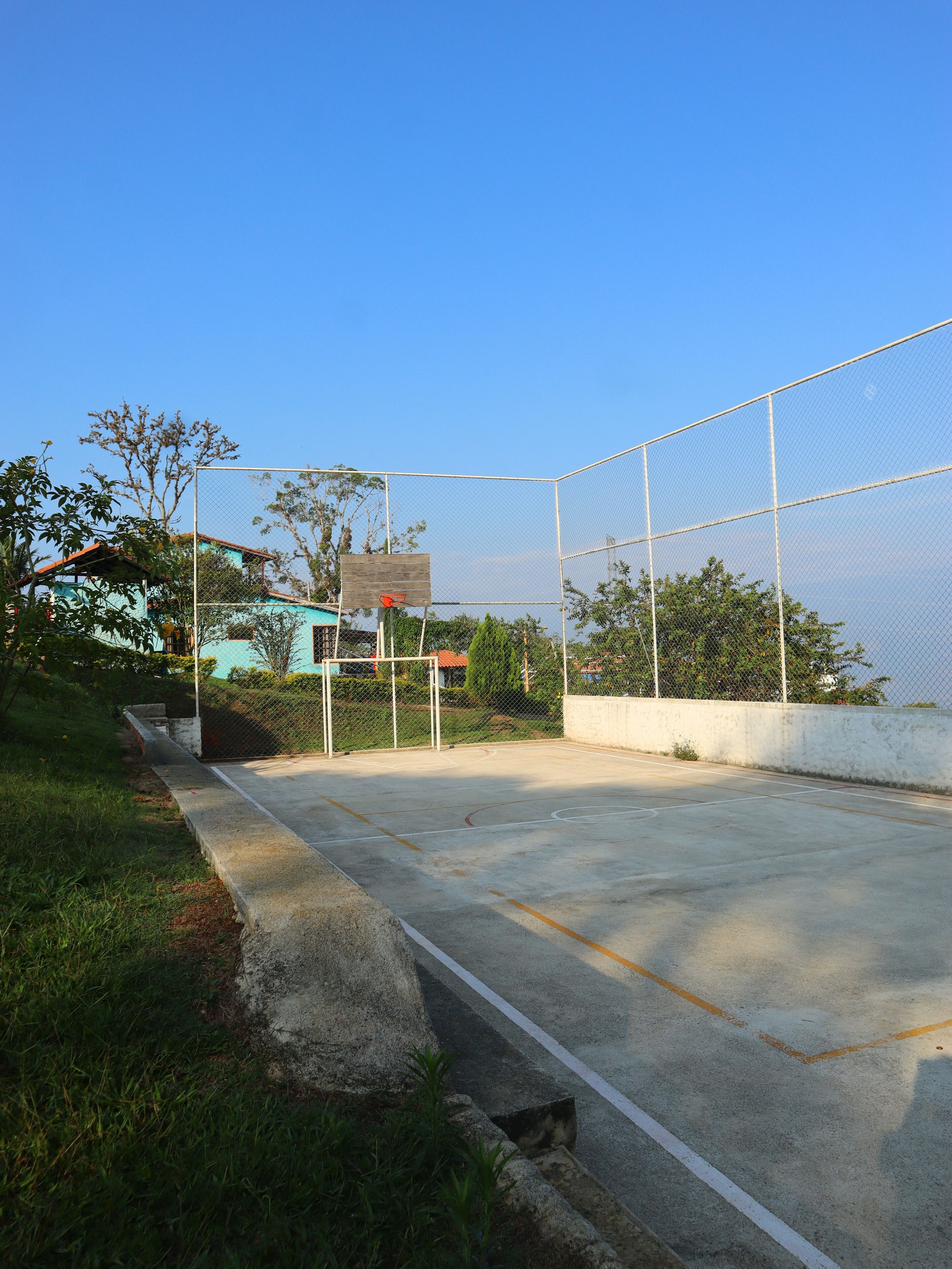 An outdoor basketball court with a net, surrounded by a chain-link fence, and a house in the background.