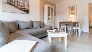 Ein modernes Wohnzimmer mit einem grauen Ecksofa, einer weißen Kommode, einem Esstisch mit Stühlen und einem Gemälde eines Leuchtturms.