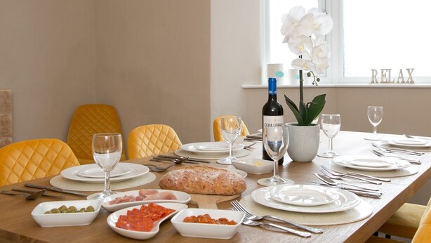 A dining table set with plates, cutlery, a bottle of wine, a loaf of bread, and various dishes including olives and cured meats. - Llys Owain, comfortable Cottage - Pass the Keys (Amlwch)