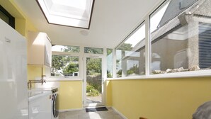 A compact kitchen with a built-in washing machine, a window with a view of greenery, and a door leading outside. - Llys Owain, comfortable Cottage - Pass the Keys (Amlwch)
