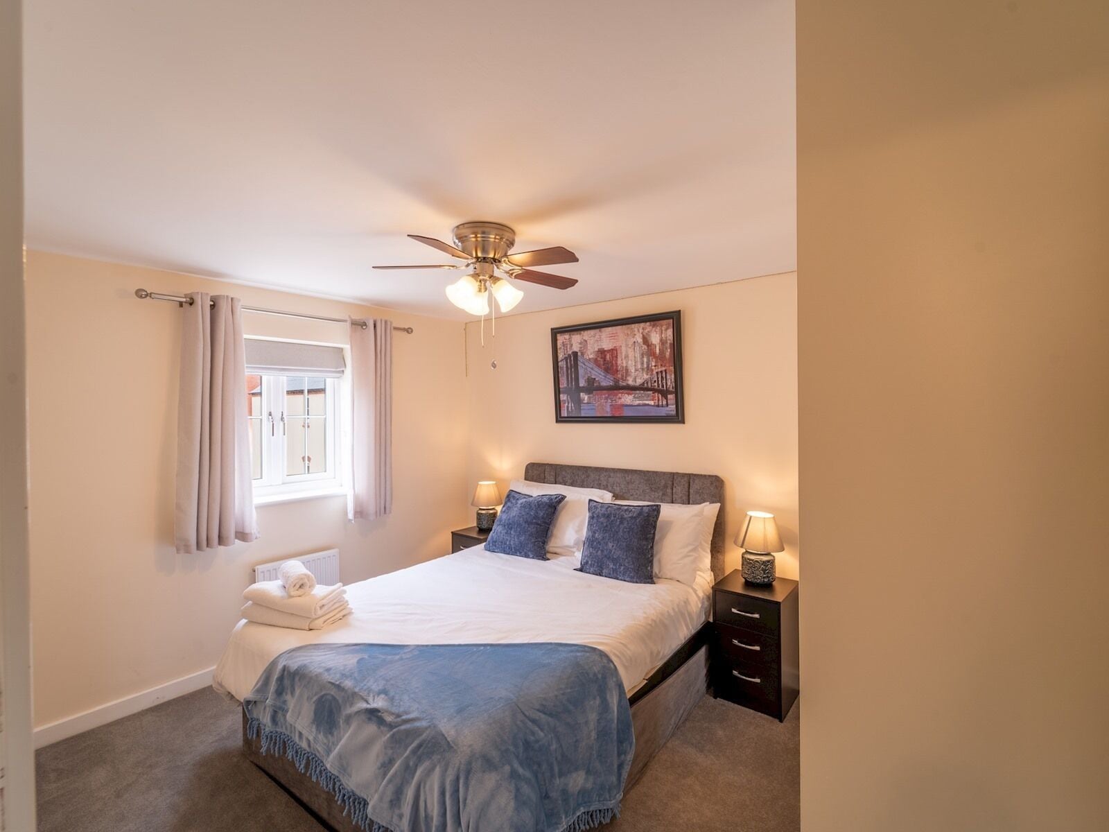 A neatly arranged bedroom with a bed, bedside tables, a ceiling fan, a window with curtains, and a painting on the wall.
