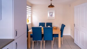 A dining area with a wooden table and blue upholstered chairs, a hanging pendant light, and a window with blinds. - Shop & Stay at Bicester Village near our 3 Bed Home-  Pass the Keys (Bicester)